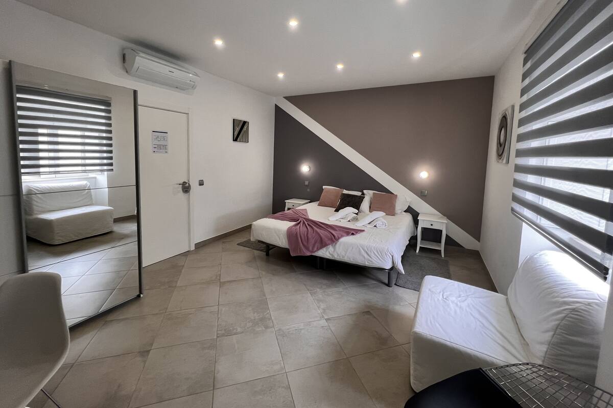 A modern bedroom with a bed, bedside tables, a chair, and a large window with blinds.