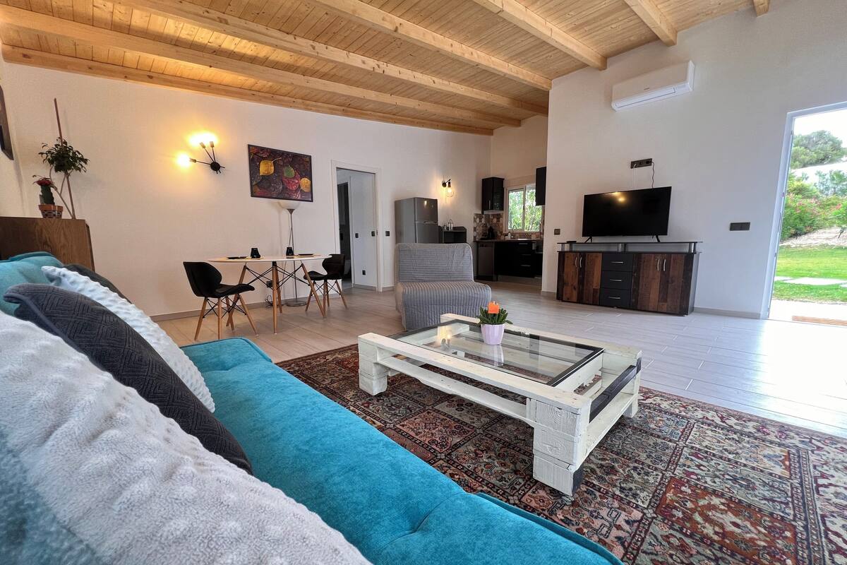 A spacious living area with a large sofa, a coffee table, and a TV. There is a dining area with chairs and a table, and a kitchen with cabinets and a TV.