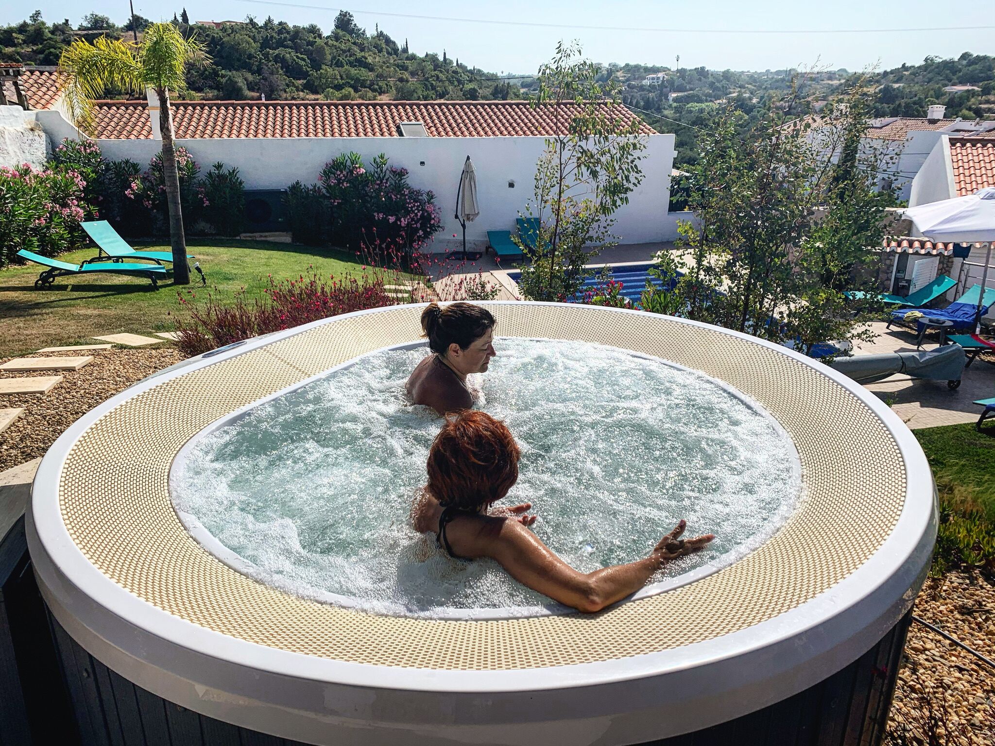 A hot tub with two people in it, surrounded by a garden with lounge chairs and a view of a hillside.
