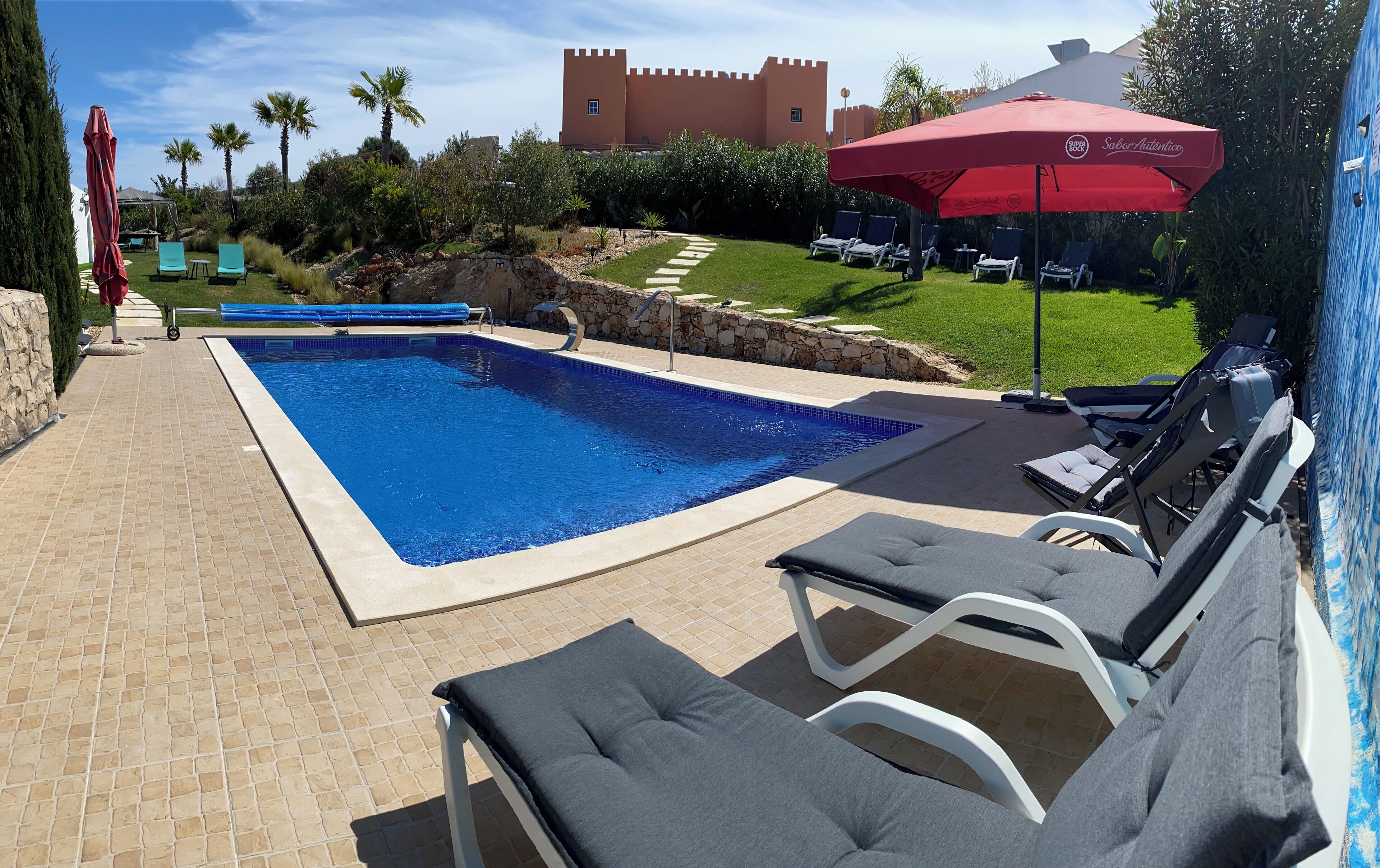 A swimming pool with lounge chairs and umbrellas, surrounded by greenery and a castle-like building.