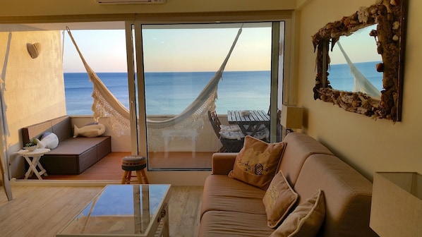 A living room with a sofa, a coffee table, and a hammock. There is a large window with a view of the ocean.