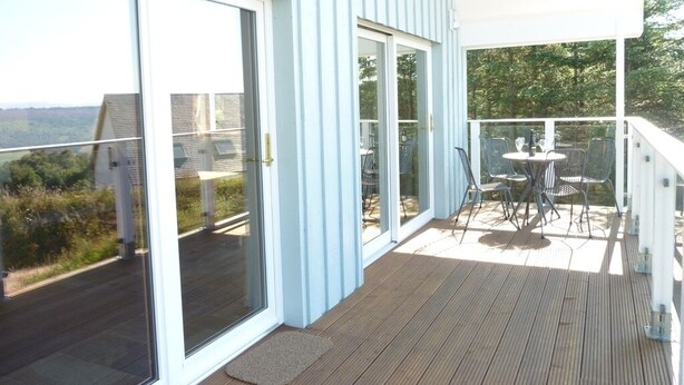 A covered outdoor area with a wooden deck, glass doors leading to a patio, and outdoor furniture. - Stylish, spacious lodge with panoramic views, an ideal base for Loch Ness (Drumnadrochit)