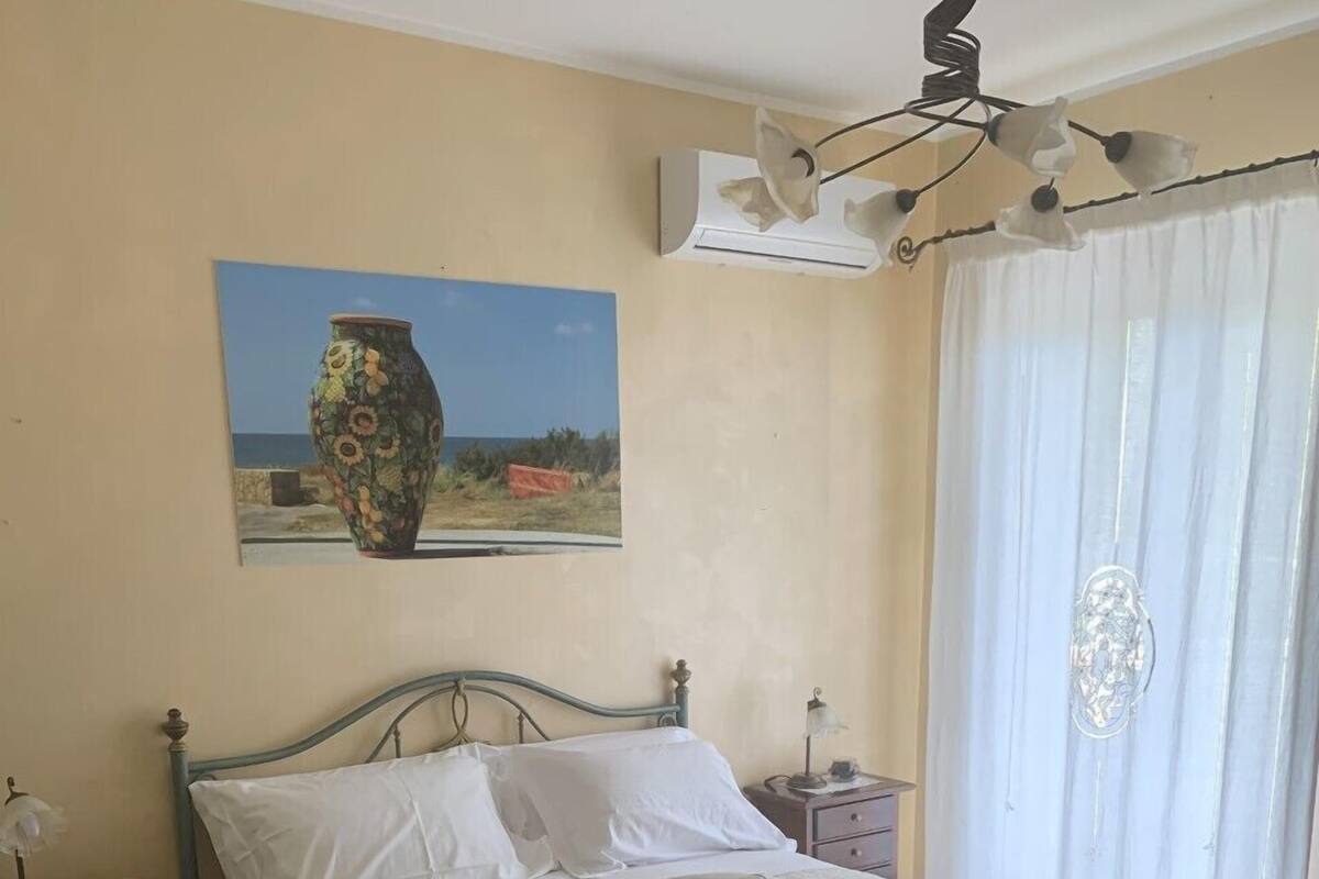 A bedroom with a bed, a nightstand, a lamp, a decorative vase on the wall, and a window with curtains.