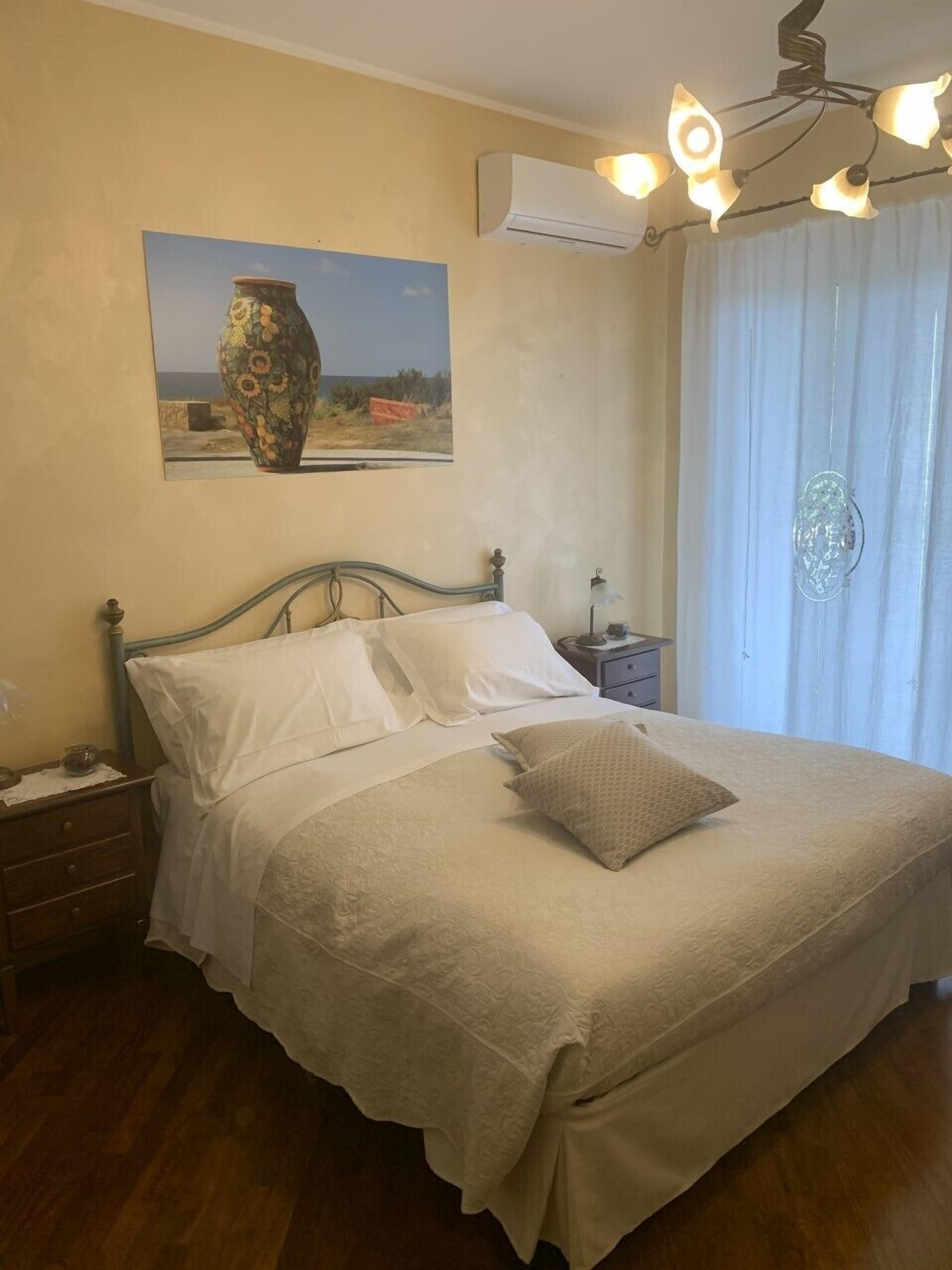 A bedroom with a bed, a nightstand, a lamp, an air conditioner, and a painting on the wall.