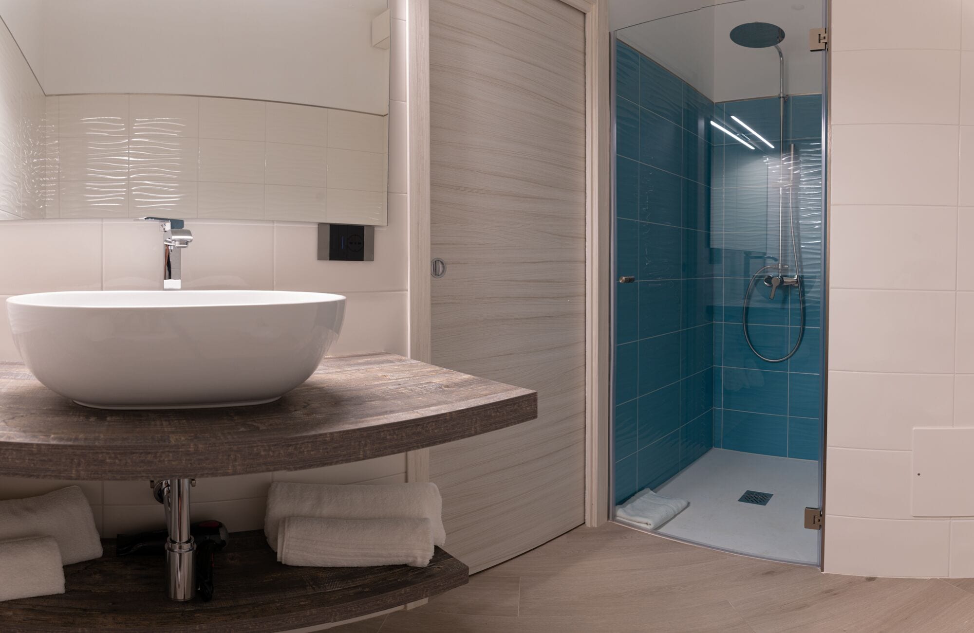 A bathroom with a large white vessel sink, a wooden countertop, a towel rack, and a glass-enclosed shower area with blue tiles.