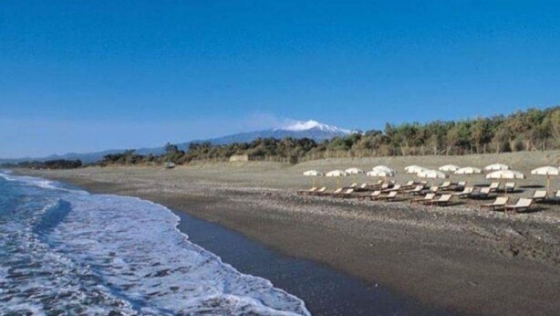 Une plage bordée de rangées de chaises longues et de parasols, une montagne en arrière-plan et un ciel dégagé.