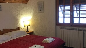 Une chambre avec un plafond en bois, une couvre-lit rouge, deux serviettes, une table de chevet avec une lampe et une fenêtre avec des stores.