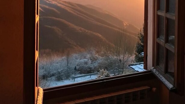 Une pièce avec une fenêtre donnant sur un paysage enneigé et des montagnes lointaines, au moment du coucher du soleil.