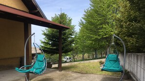 Deux chaises à bascule vertes se trouvent sous une terrasse couverte, dont le sol est en béton. L’ensemble est entouré d’arbres et d’une clôture.