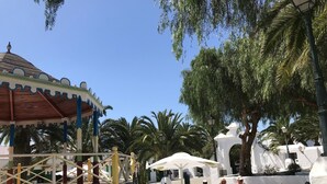 A sunny outdoor seating area with wicker chairs and umbrellas, surrounded by palm trees and a white building with a dome roof.