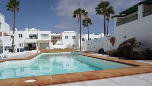 A swimming pool with a tiled edge, surrounded by white buildings, palm trees, and a rocky wall.