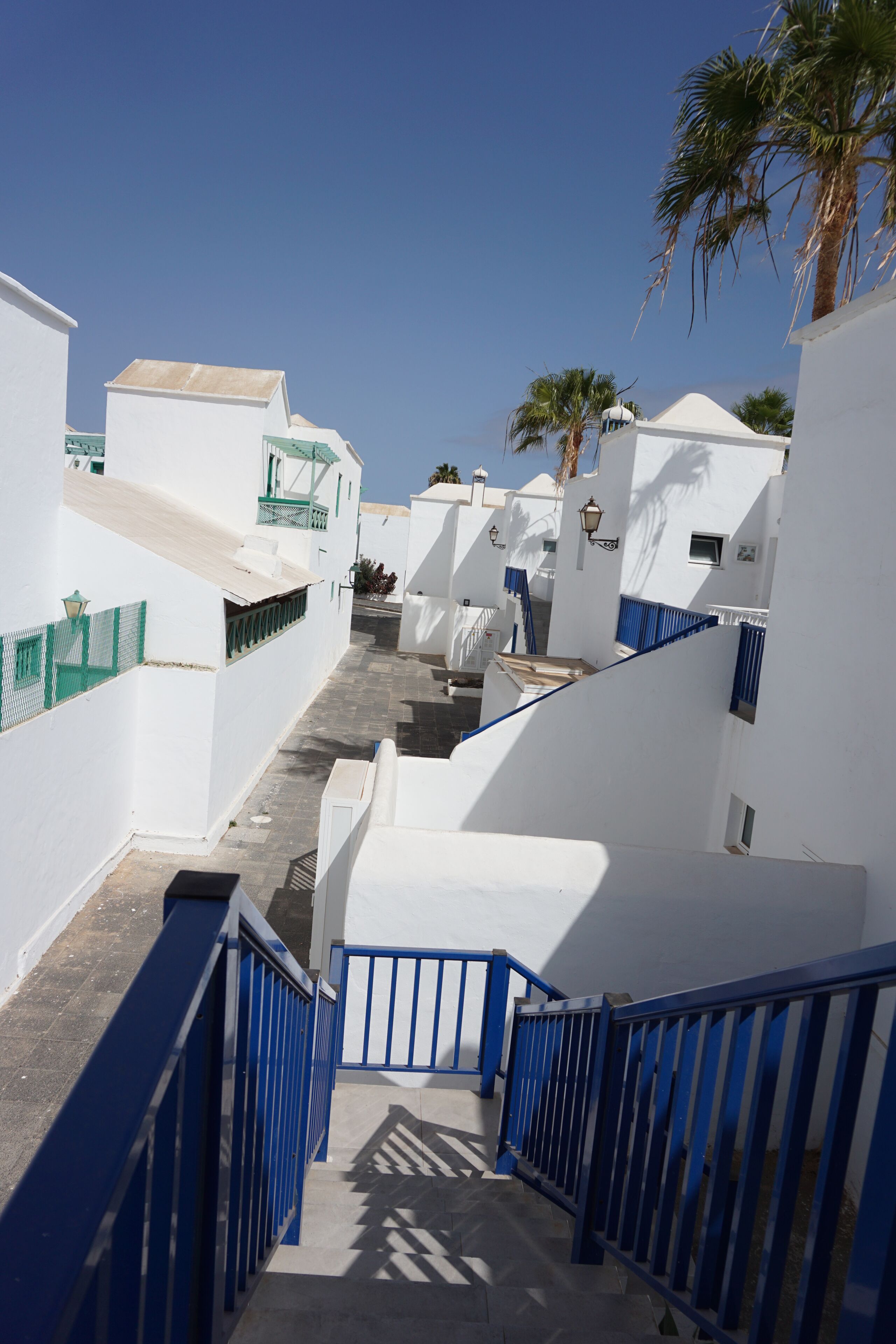 A narrow, sunlit street with white buildings and blue railings, leading to a white building with a blue door.
