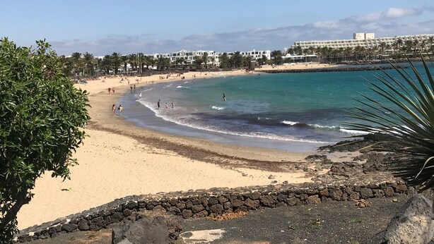 A beach with a stone wall, clear blue water, and buildings in the background.