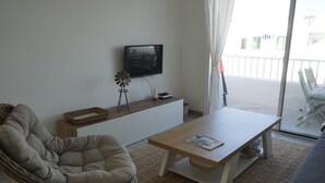 A living room with a sofa, a wicker chair, a coffee table, a TV on a wall, a small windmill decoration, and a balcony with a view.