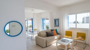 A modern living room with a sofa, a dining table, and yellow chairs. There is a large window with a view of buildings and a door leading to another room.
