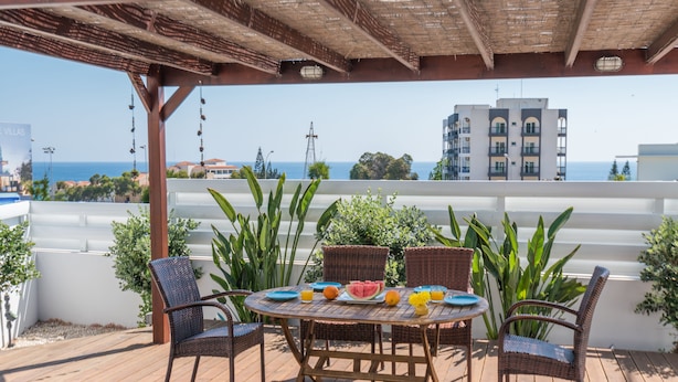 A covered outdoor dining area with wicker furniture and a table set with fruits and drinks, overlooking a coastal cityscape.