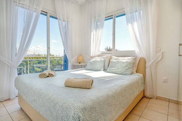 A bedroom with a large bed, white curtains, and a view of greenery outside.