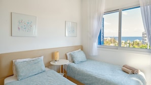 A hotel room with two beds, a small table, and a window with a view of the sea and buildings.