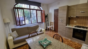 Un ambiente compatto con divano, tavolo da pranzo, angolo cottura e finestra vista su piante.