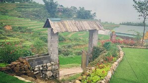 Un arco de piedra rústico que da paso a un jardín con un estanque, rodeado de campos escalonados y un cielo despejado.