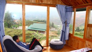 Habitación con un amplio ventanal que ofrece vistas a campos escalonados y una casa. Se observa un sillón informal y un pequeño banquillo redondo.