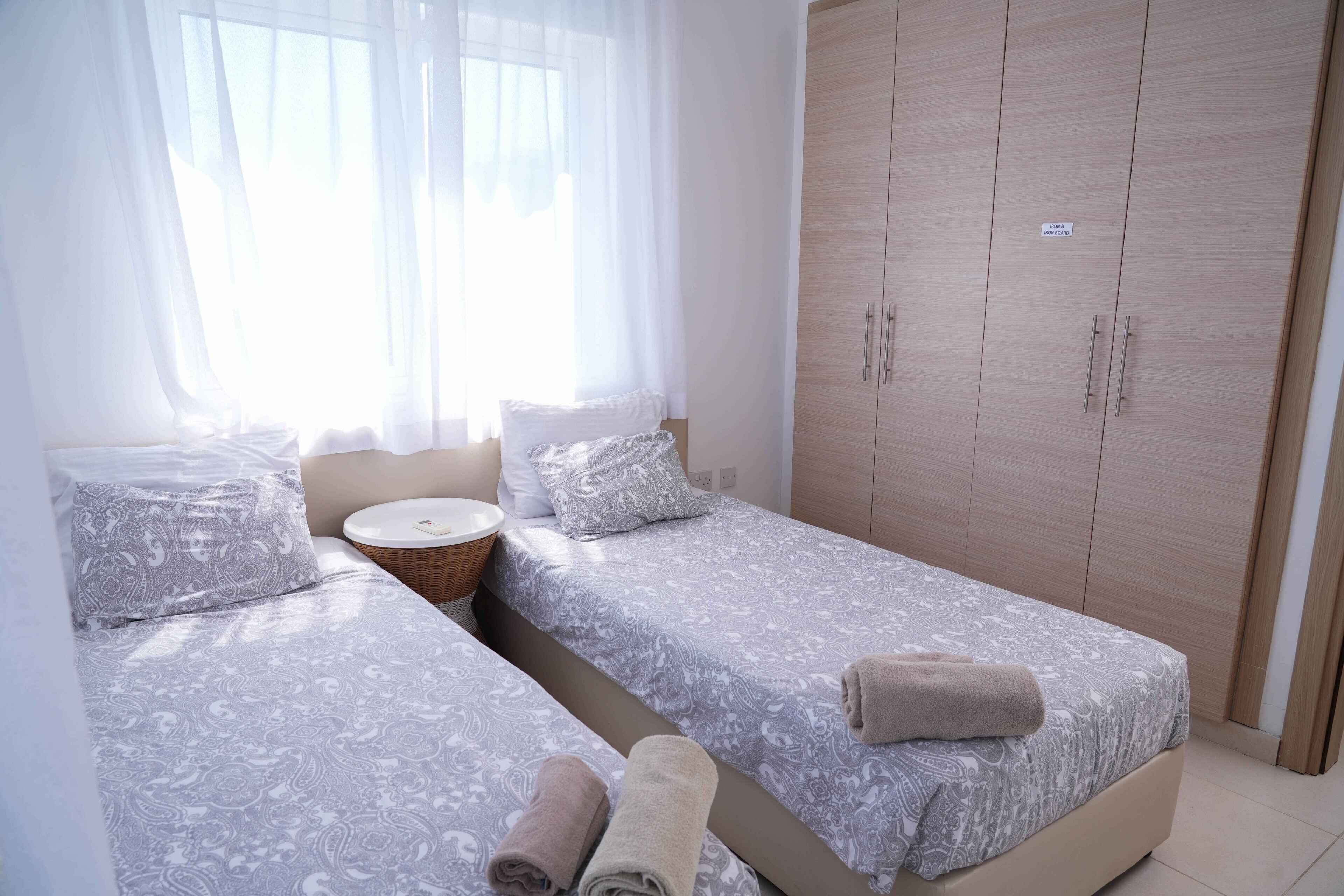 A bedroom with two beds, a wooden wardrobe, and a bedside table with a basket.