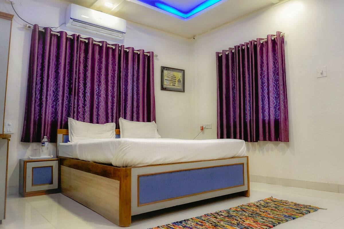 Une chambre à coucher avec un lit, des rideaux violets, une petite table de chevet, une commode, un climatiseur fixé au mur et une porte en bois.
