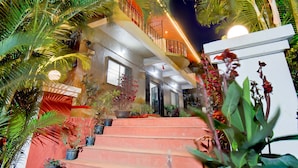 Un immeuble à plusieurs étages, doté d’un escalier menant à un balcon, lequel est entouré de plantes en pot et de palmiers.