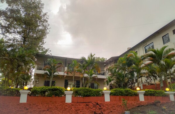 Un edificio de varios pisos con balcones, rodeado de abundante vegetación y un muro rojo.