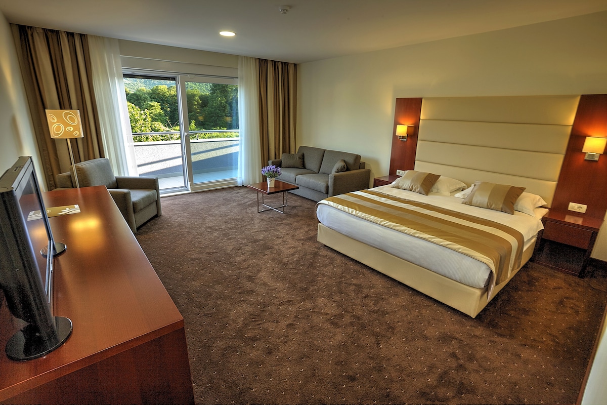 Habitación de hotel con una cama grande, un sofá, un escritorio y vistas a zonas verdes a través de las ventanas.