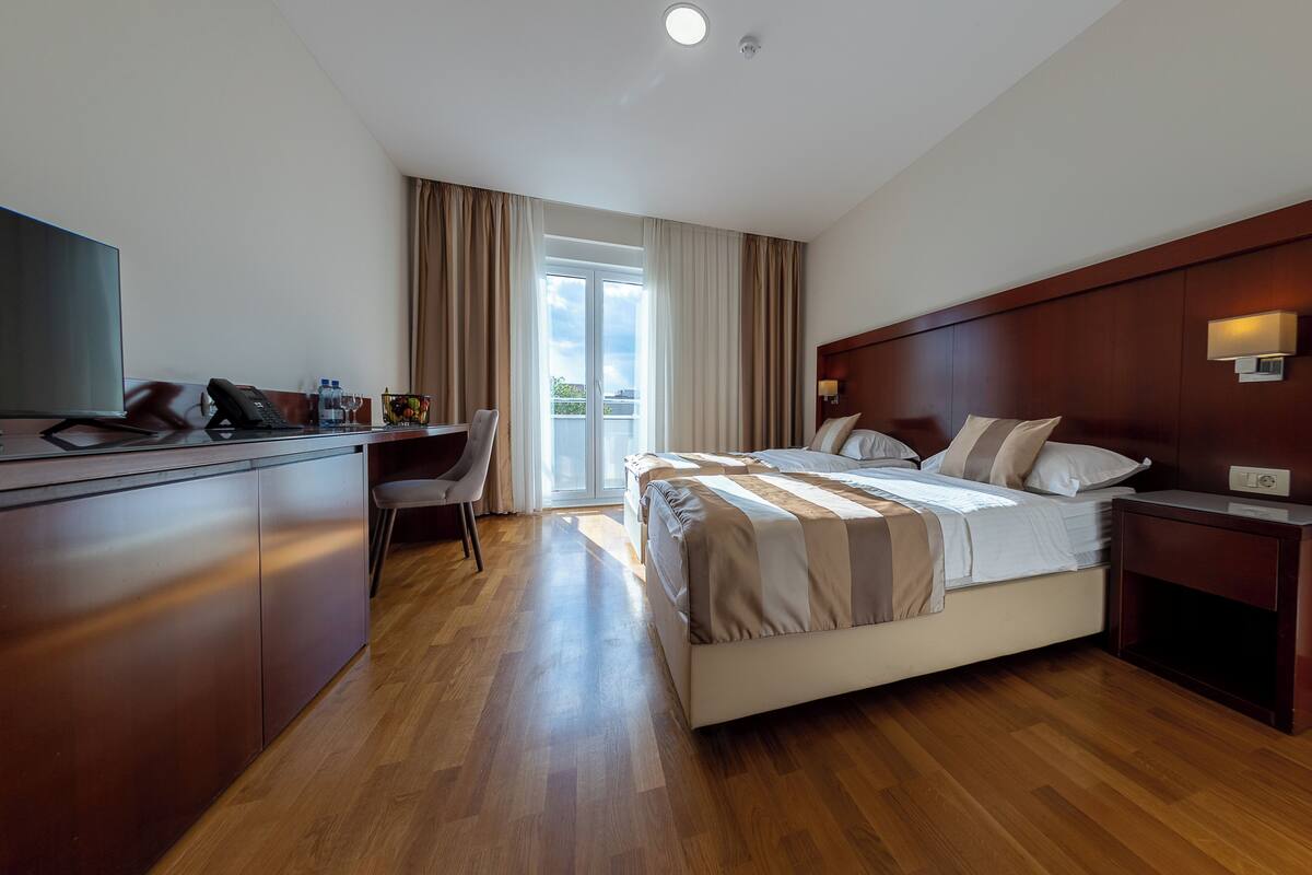 A hotel room with a desk, two beds, a TV, and a view of the outdoors.