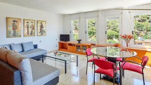 Uma sala de estar moderna com um sofá, uma mesa de centro de vidro e uma poltrona vermelha dispostas ao redor de uma mesa de jantar de vidro. Há obras de arte emolduradas na parede e uma TV sobre um suporte de madeira.