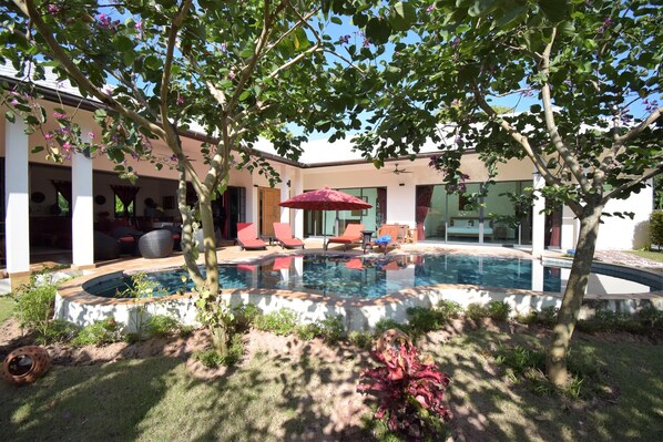 A modern outdoor pool area with a hot tub, surrounded by a covered patio with red chairs and a red umbrella.