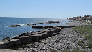 Ein felsiger Strand mit blauem Himmel, ruhiger See und einer Reihe großer Betonblöcke.