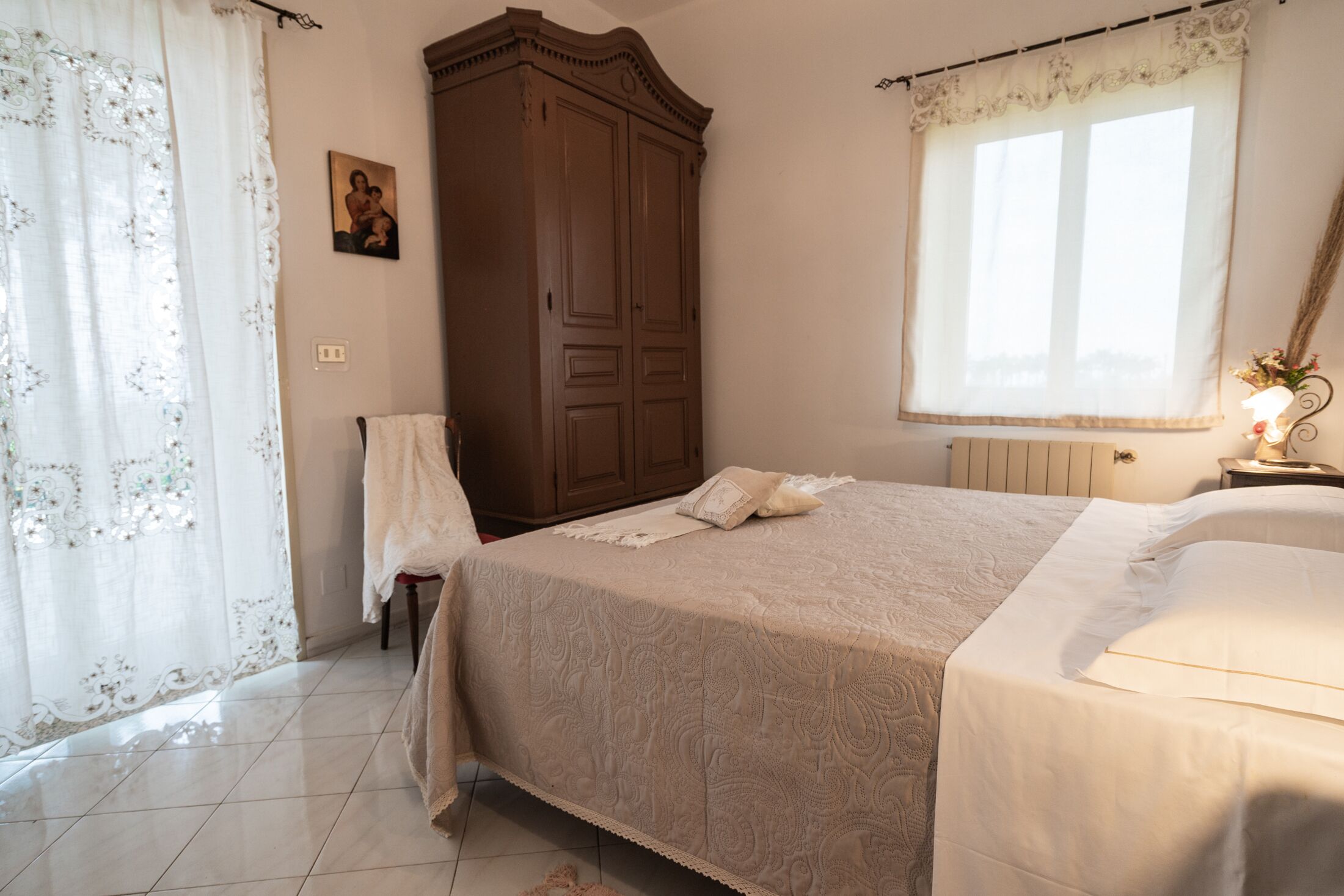 A bedroom with a bed, a wardrobe, a window with curtains, and a nightstand with a lamp.