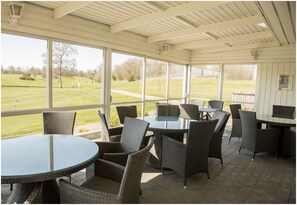 Een overdekt terras met rieten meubilair en grote ramen met uitzicht op een golfbaan.