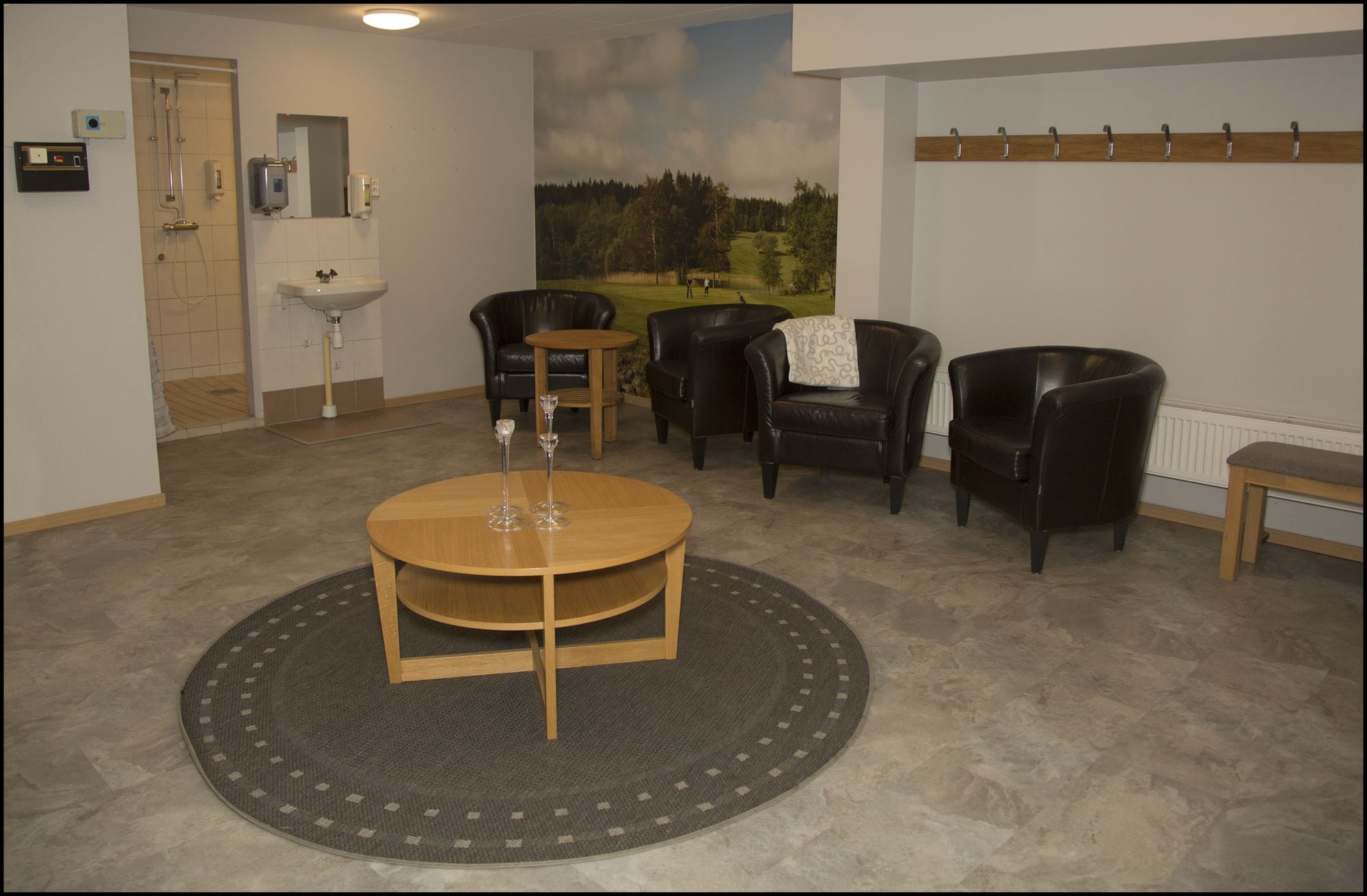 A room with a round wooden table, four black leather chairs, a small round table with two wine glasses, a shower area, a sink, and a wall-mounted towel rack.