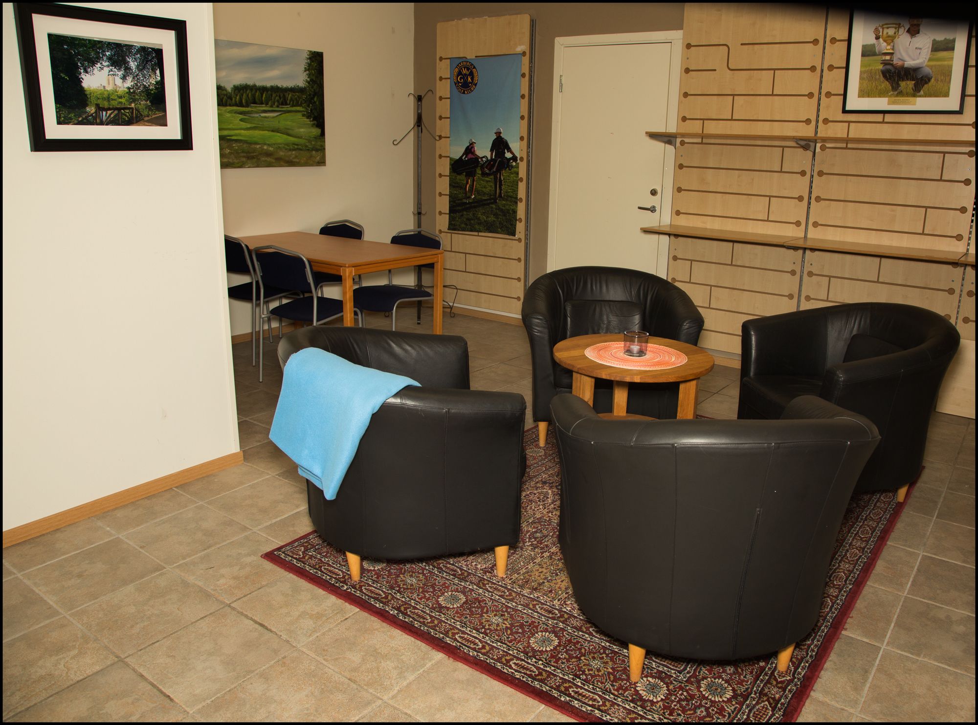 A living room with black leather sofas, a wooden coffee table, and a red patterned rug. There is a door with a poster and a framed picture on the wall.