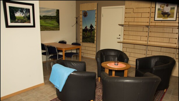 Un salon avec des canapés en cuir noir, une table basse en bois et un tapis à motifs rouges. On y trouve une porte ornée d’une affiche et un tableau encadré accroché au mur.