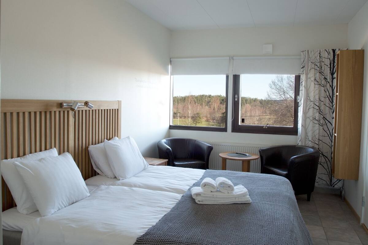 Une chambre d’hôtel avec un lit, deux chaises, une petite table et une fenêtre donnant sur des arbres.