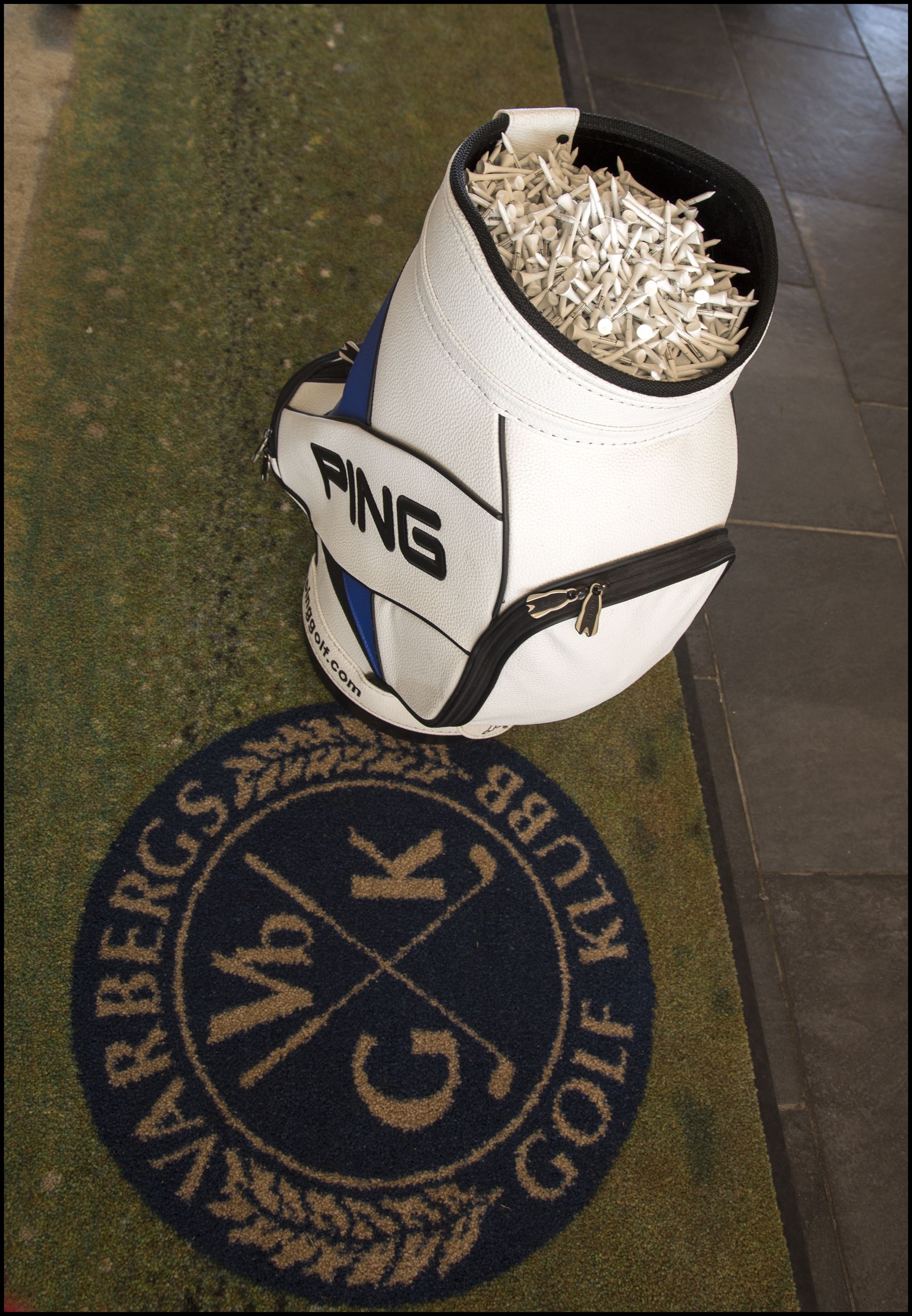 Un sac de golf de la marque « PING » est posé sur un tapis orné du logo « MERRICK’S GOLF CLUB ».