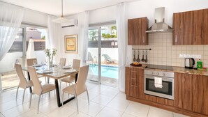 A modern kitchen with wooden cabinets, a built-in oven, and a dining area with a glass table and chairs. - Narcissos Villa 2 (Paralimni)