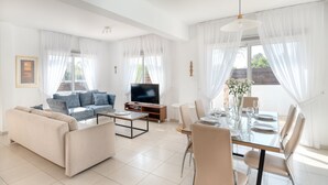 A modern living room with a sofa, TV, coffee table, and dining area with chairs and a table set for four. - Narcissos Villa 2 (Paralimni)