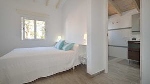 Un dormitorio con cama, almohadas, mesita de noche, ventana y baño con lavabo y espejo.