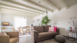 Uma sala de estar com sofá, uma poltrona, mesa de jantar com cadeiras, planta e uma pequena mesa lateral.