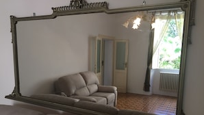 Een kamer met een grote spiegel die een beige fauteuil, een deur en een radiator weerspiegelt.