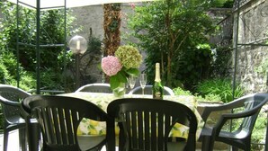 Een terras buiten met een ronde tafel, zwarte plastic stoelen, een vaas met bloemen en een fles champagne.