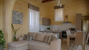 A cozy living room with a beige sofa, a dining table, and a kitchen area with a stove and cabinets. - Simar Farmhouse (Qala)