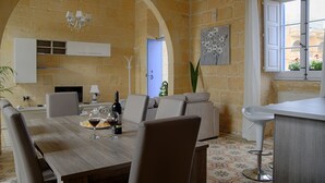 A dining area with a wooden table, chairs, and a chandelier. There is a blue door leading to another room, and a window with curtains. - Simar Farmhouse (Qala)