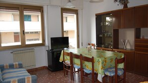 A dining area with a table set for four, a television, and a window with blinds. - VERSILIA APARTMENT (Pietrasanta)